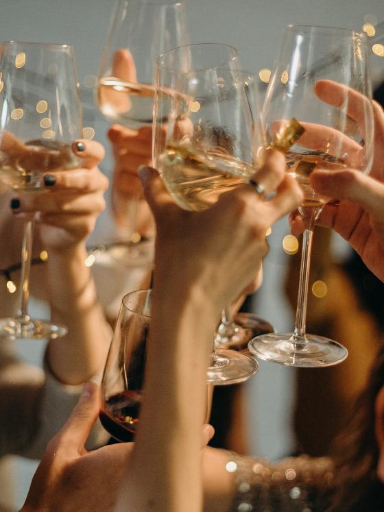 Image of a holiday party and people clinking glasses of white wine, saying "cheers".