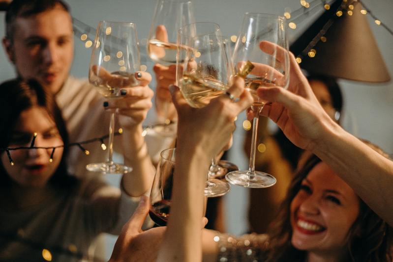Image of a holiday party and people clinking glasses of white wine, saying "cheers".
